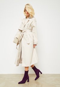 Trench beige con cinturón, botones de doble botonadura y amplia solapa. Complementa unas botas de tobillo violetas con tacón de bloque y puntera afilada.