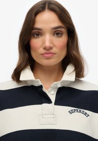 Niebiesko-kremowa w paski koszulka polo z zapięciem na guziki przy kołnierzyku, z haftowanym logo "Superdry" na klatce piersiowej. Wykonana z bawełny, luźny krój.