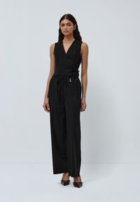 Svart ermeløs jumpsuit med innpakningsdesign, innsnevret midje, avslappede ben, subtil glans og dekorativ snor detalj.
