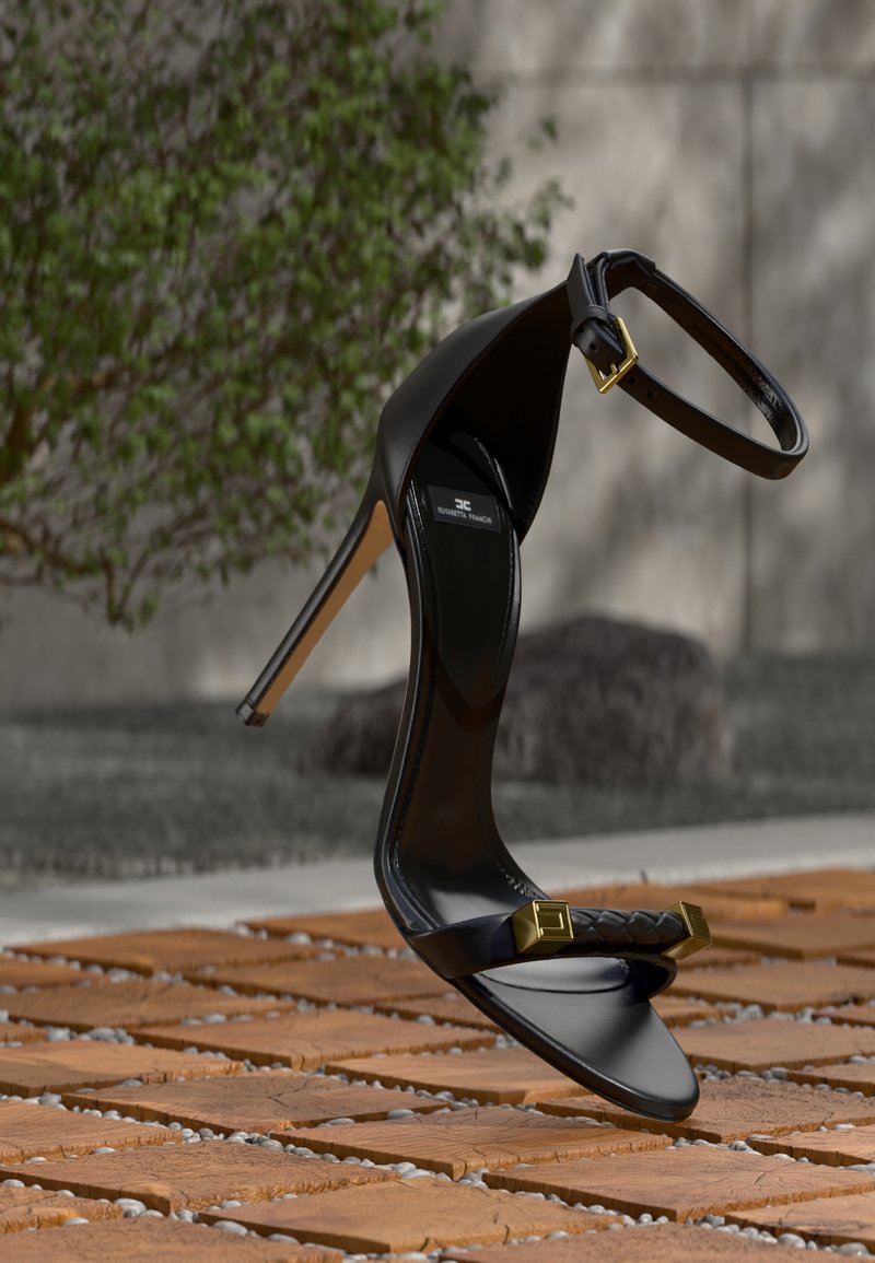 Μαύρο πέδιλο υψηλού τακουνιού με λουρί στον αστράγαλο και χρυσή αγκράφα, πλεκτό μπροστινό λουρί με χρυσές λεπτομέρειες, αιωρούμενο πάνω από ξύλινο και πλακόστρωτο δάπεδο.