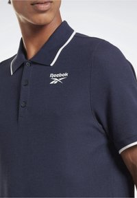 Námořnická modrá polo košile s texturovanou tkaninou, bílým límcem a logem Reebok na levém prsou. Obsahuje lištu na tři knoflíky.