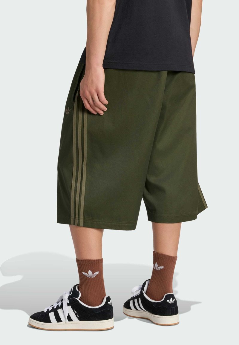 Persona que lleva pantalones cortos color verde oliva hasta la rodilla con rayas laterales, calcetines marrones de Adidas, zapatillas negras de Adidas, de pie sobre un fondo claro.
