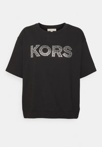 Camisa de manga corta negra con cuello redondo, que presenta "KORS" escrito en tachuelas plateadas en el pecho.