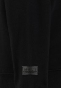 Schwarzer Pullover mit glatter Textur. Verfügt über ein auffälliges schwarzes Etikett an der unteren linken Seite mit erhabenem Text. Lange Ärmel, gerippte Bündchen.