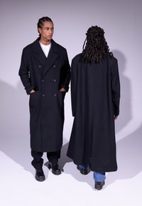 Manteau long en laine noir avec un devant croisé, des revers larges, des poches latérales et un tissu texturé. Présenté de face et de dos.