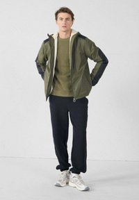 Olijfgroene jas met een fleece gevoerde kraag, zwarte accenten en ritssluiting, gecombineerd met een groene trui en zwarte sweatpants.