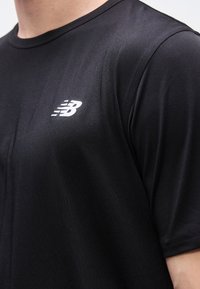 Μαύρο αθλητικό μπλουζάκι με λευκό λογότυπο New Balance στο πάνω μέρος του στήθους και της περιοχής του ώμου ενός ατόμου.