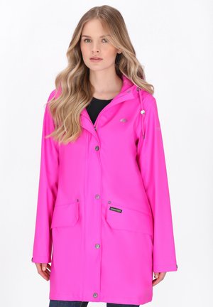 Donna con capelli lunghi e biondi che indossa un impermeabile rosa brillante con tasche anteriori e bottoni a pressione, in piedi davanti a uno sfondo bianco.