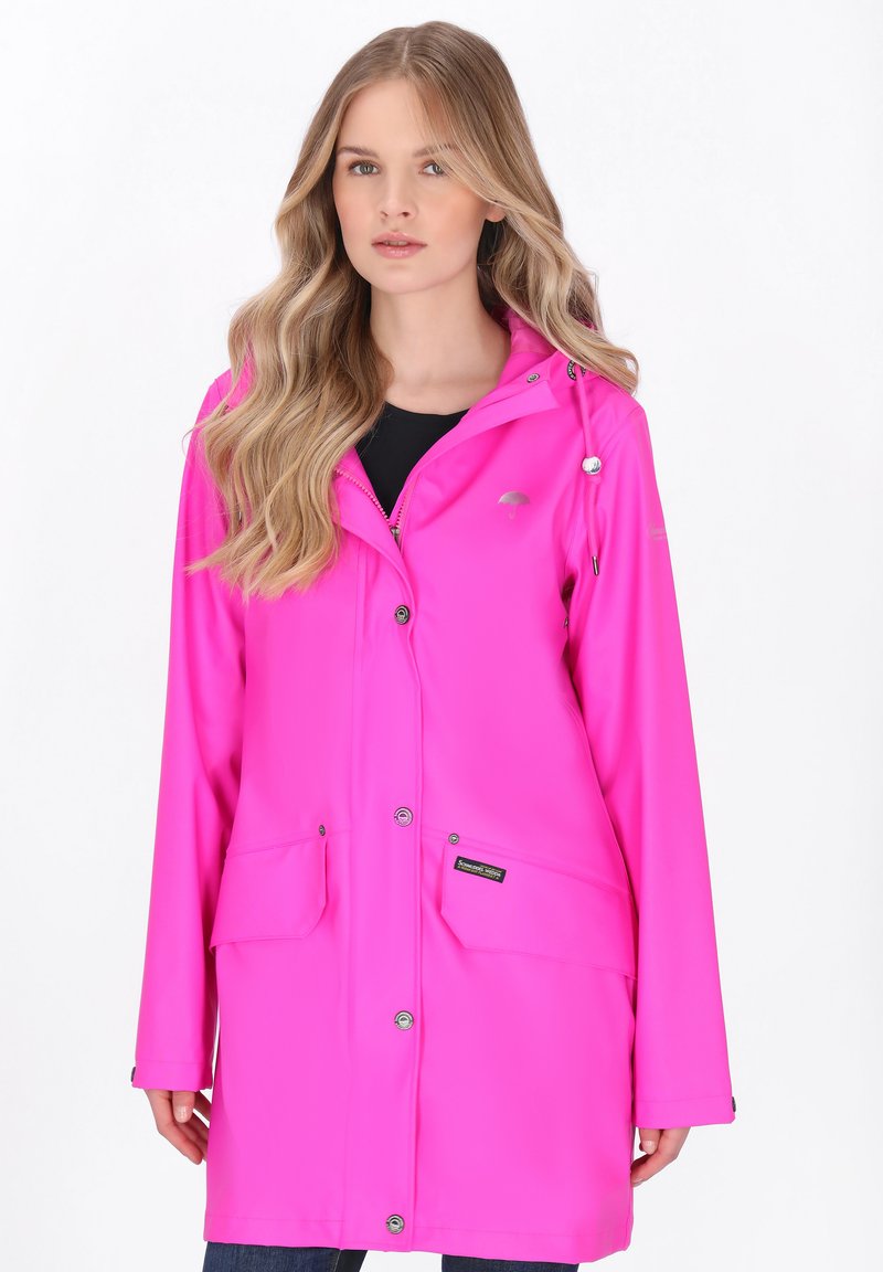 Donna con capelli lunghi e biondi che indossa un impermeabile rosa brillante con tasche anteriori e bottoni a pressione, in piedi davanti a uno sfondo bianco.