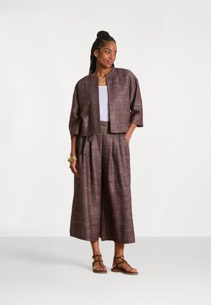Femme aux cheveux tressés, portant une veste courte marron texturée et un pantalon large, un top blanc, des sandales et un bracelet en or, sur fond blanc.