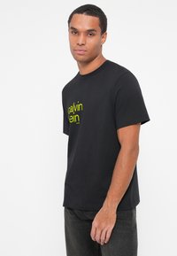 Juoda medvilninė marškinėliai su trumpomis rankovėmis, kuriuose ant priekinės dalies yra ryški geltona "Calvin Klein" logotipo dizainas. Klasikinis siluetas ir apvalus kaklas.