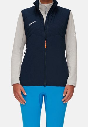 Person trägt eine dunkelblaue ärmellose Weste über einem hellgrauen Langarmshirt mit leuchtend blauen enganliegenden Hosen, steht mit entspannten Händen.