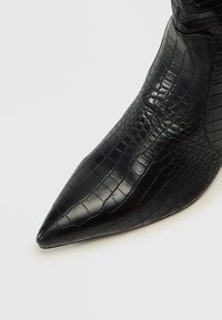 Botín negro de punta afilada hecho de cuero con textura de cocodrilo brillante; presenta una textura suave y un diseño elegante.