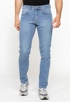 Uomo che indossa jeans slim fit azzurri chiaro e sneakers grigie, in piedi davanti a uno sfondo bianco semplice.