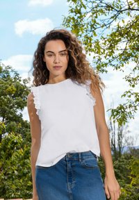 Top bianco senza maniche con rifinitura in pizzo arricciato sulle spalle, abbinato a una gonna di jeans blu. Lo sfondo presenta vegetazione verde e cielo blu.