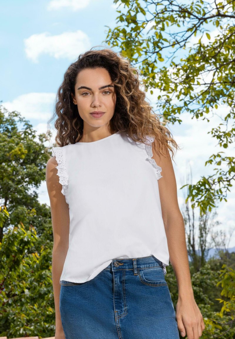 Top bianco senza maniche con rifinitura in pizzo arricciato sulle spalle, abbinato a una gonna di jeans blu. Lo sfondo presenta vegetazione verde e cielo blu.