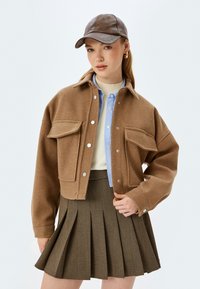 Brązowa, wełniana kurtka w kroju cropped z dwoma przednimi kieszeniami i srebrnymi zatrzaskami, w połączeniu z jasnoniebieską koszulą zapinaną na guziki oraz plisowaną brązową spódnicą.