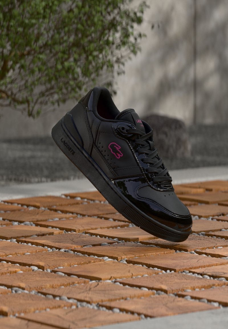 Baskets noires à finition brillante et logo Lacoste rose semblant flotter au-dessus d’un sol en carrelage à motifs, avec des arbres et un mur en béton en arrière-plan.