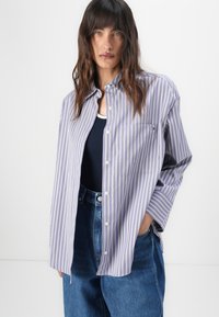Camicia a righe in morbido viola e bianco, con taschino sul petto e maniche arrotolate, indossata sopra un top in navy.