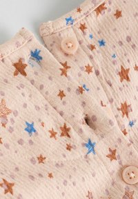 Vêtement en coton de couleur pêche claire, orné de motifs d'étoiles bruns et bleus, et avec deux boutons rose clair près de l'encolure.