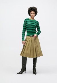 Πλεκτό πουλόβερ με πράσινες και ναυτικές ρίγες, με ριμπ υφή, συνδυασμένο με πλισέ μπεζ φούστα και μαύρες δερμάτινες μπότες μέχρι το γόνατο.