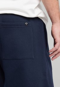 Sotkansiniset jogger-housut, jotka on valmistettu pehmeästä materiaalista. Niissä on takana läpällä varustettu tasku ja pieni logo-merkki vyötärön lähellä.