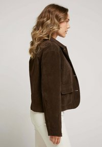Femme avec des cheveux blonds ondulés portant une veste en daim marron foncé et un pantalon blanc, faisant face à droite sur un fond uni.