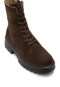 Stivale alla caviglia in suede marrone con design lacciato, colletto imbottito e suola in gomma spessa con scanalature orizzontali. Lacci e dettagli di cucitura marroni.