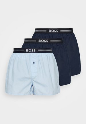 Szczupły, muskularny mężczyzna bez koszulki stoi boso, ma na sobie jasnoniebieskie bokserki z czarną gumą w pasie z napisem „BOSS” na jednolitym białym tle.