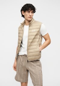 Colete acolchoado bege com design acolchoado vertical, gola alta e logótipo EA7. Usado sobre uma t-shirt branca e combinado com calções bege estampados.