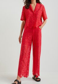 Haut rouge en dentelle avec des boutons, à manches courtes et un pantalon large assorti, présentant des motifs complexes et des ourlets festonnés, porté avec des sandales noires.