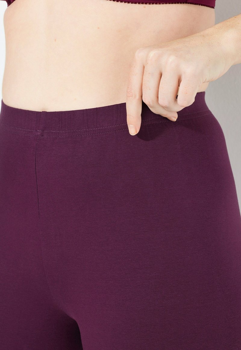 Leggings morados hechos de un tejido suave y elástico con una cinturilla ancha. Un dedo señala la cinturilla, destacando su detalle de diseño.