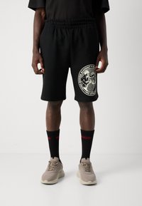 Personen iført sorte shorts med hvit boksing-grafikk, svarte sokker med røde tekster, og beige sneakers på hvit bakgrunn.