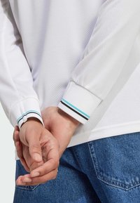 Bijela teksturirana košulja dugih rukava s elastičnim manžetama u tirkiznim i crnim prugama, u kombinaciji s trapericama. Ruke spojene iza leđa.
