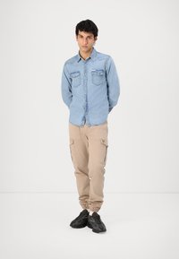 Camisa de mezclilla azul claro con dos bolsillos en el pecho, combinada con pantalones cargo beiges con puños elásticos y bolsillos laterales, y zapatillas negras.