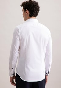 Camicia bianca a maniche lunghe con colletto a punta, dotata di polsini con bottoni e accenti blu navy e orlo curvo. Tessuto liscia.