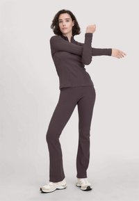 Frau in dunkelbrownem Sportoutfit, die ihren rechten Arm über die Brust dehnt, trägt weiße und beige Sneakers vor einem einfarbigen Hintergrund.