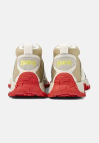 Baskets beiges et blanches avec une semelle extérieure en caoutchouc rouge, dotées d'un talon texturé, d'une languette et d'un logo 'Camper' en relief en jaune sur le talon.