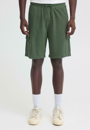 Mężczyzna w zielonych spodniach typu cargo, białych skarpetach za kostkę i kremowych trampkach, stoi na tle jednolitego tła.