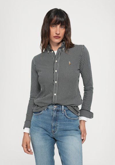 Chemise à rayures noires et blanches avec col, arborant un petit logo brodé et des manches retroussées, associée à un jean taille haute bleu.
