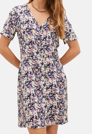 Woman wearing a short-sleeve, button-front dress with a multicolor floral pattern and a gold chain necklace.