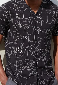 Zwarte korte mouwen overhemd met knopen, bedekt met witte abstracte gezichten en botanische ontwerpen in één doorlopende lijn, gedragen door een persoon met de handen in de zakken.