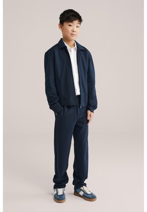 Jongen staand, gekleed in een marineblauwe jas met rits, bijpassende broek, wit overhemd met kraag en blauwe sneakers tegen een effen achtergrond.