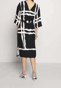 Abito nero con strisce astratte bianche, design a V sulla schiena, maniche ampie e uno spacco laterale, abbinato a scarpe con tacco alto bianche e con cinturini.