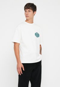 White cotton t-shirt with a round neckline and short sleeves, featuring a teal badge graphic on the chest that says "Notes of Nostalgia".