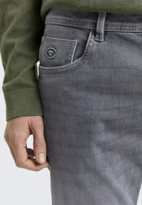 Szare dżinsy z denimu mają z przodu kieszeń z okrągłym logo, w połączeniu z zielonym dzianinowym swetrem. Tekstura wydaje się gładka i dopasowana.