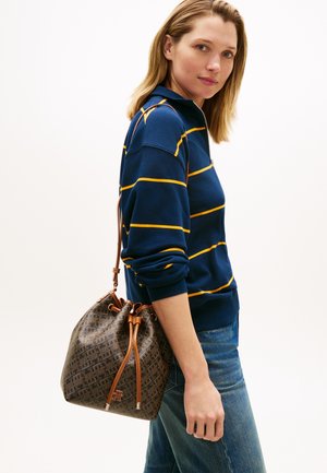 Vrouw die een blauw-geel gestreept shirt met lange mouwen en een spijkerbroek draagt, met een bruine leren emmerzak met patroon over haar schouder.