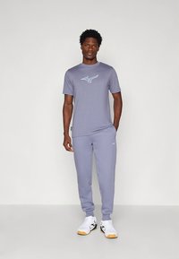 Pilkas sportinis marškinėlis su mėlynu logotipu, derinamas su atitinkančiomis laisvalaikio kelnėmis. Audinys atrodo lygus ir prigludęs, papildytas baltais sportbačiais.