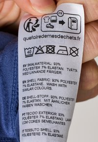Etykieta z instrukcjami dotyczącymi pielęgnacji odzieży, w przeważającej części biała z czarnym tekstem w wielu językach, zawierająca symbole prania i skład materiału.