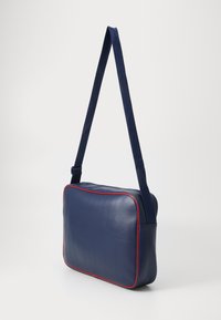 Saco de couro azul-marinho a tiracolo com acabamento liso, detalhes em vermelho e uma alça de tecido. Forma retangular com fecho com zíper e sem hardware visível.